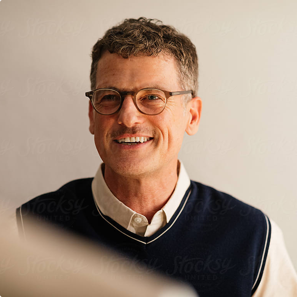 Man wearing glasses and a navy sweater over a white shirt against a plain background