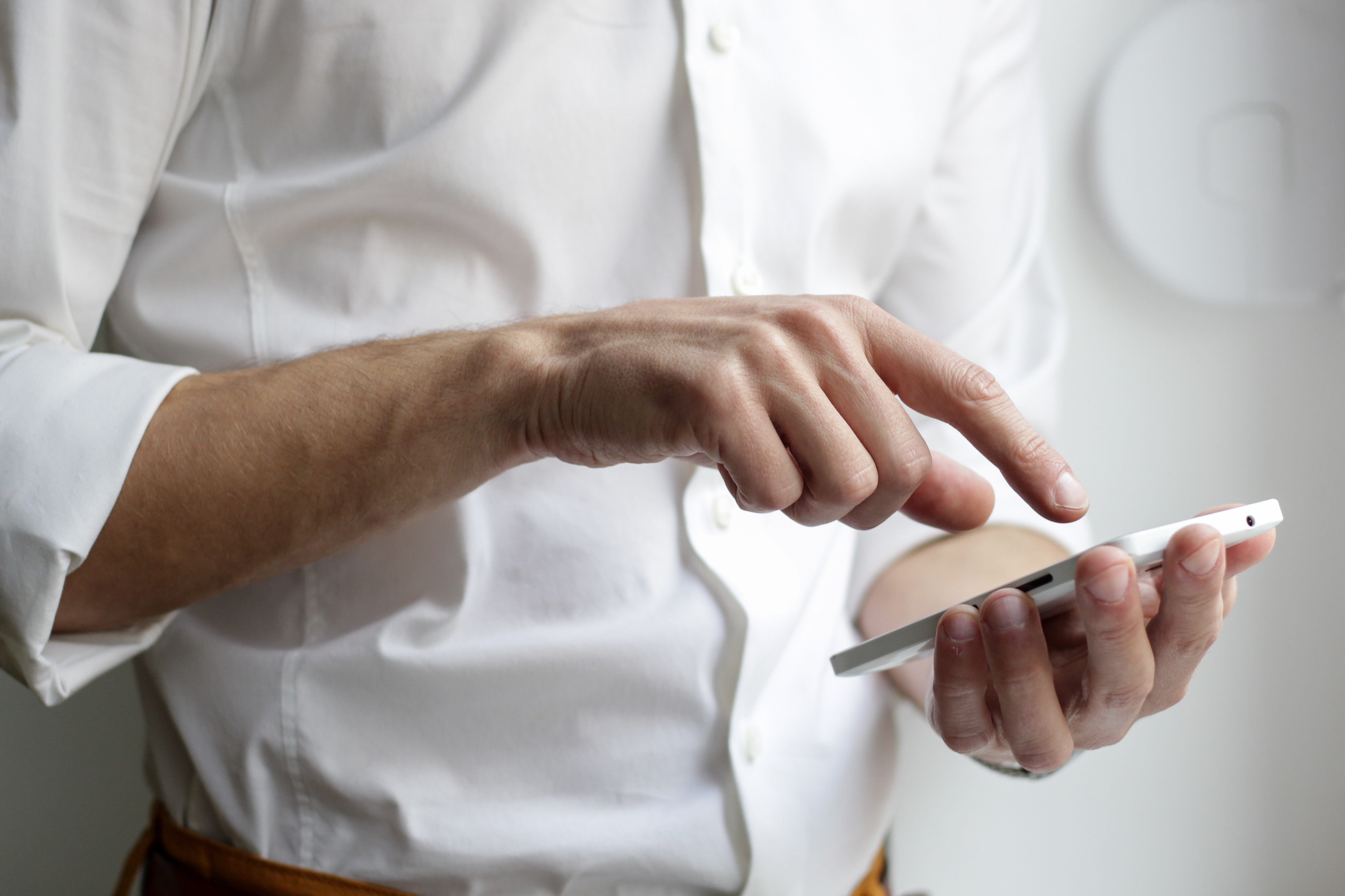 person in white button-down dress shirt holds cell phone with left phone while using index finger of right hand on screen